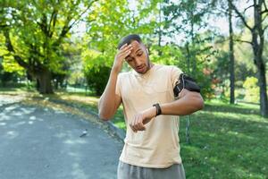 giovane africano americano uomo corridore, atleta nel il parco. un' stanco uomo conta il suo pulse su un' fitness braccialetto. sorpreso, lui sembra a il risultato su il orologio intelligente su il suo mano. salviette sudore con il suo mano. foto