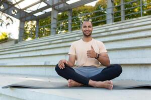 giovane sportivo nel loto posa Meditare e guardare a telecamera, con gioia mostrando pollici su, soddisfatto fitness allenatore su esercizio stuoia a stadio. foto