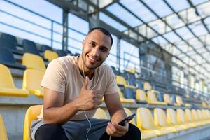 ritratto di un' giovane africano americano maschio atleta, allenatore seduta nel un' stadio indossare cuffie e utilizzando il Telefono. lui punti a il telecamera con un' super dito, sorrisi, riposa. foto