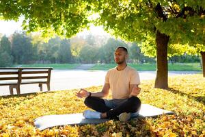 un' giovane latino americano uomo è fare yoga nel il mattina nel il parco. seduta su un' stuoia nel il loto posizione con chiuso occhi e meditando. foto