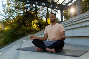 un' giovane africano americano uomo è seduta su un' stuoia nel un' stadio nel il loto posizione e fare yoga. lui medita, chiuso il suo occhi, rilassato, calma. foto