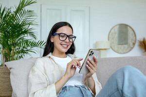 un' giovane donna è dire bugie su il divano a casa e sorridendo utilizzando un' mobile Telefono. foto