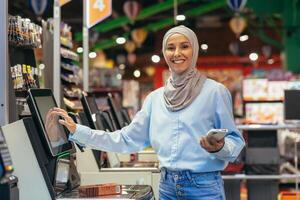 Fai da te nel un' supermercato. un' giovane arabo donna nel un' hijab è in piedi vicino il elettronico denaro contante registri e è pagare per il merce nel il memorizzare in linea attraverso un applicazione su sua Telefono. foto