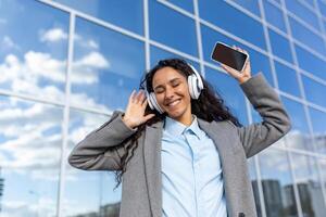 giovane bellissimo latino americano donna passeggiate nel il città, donna nel cuffie ascolta per musica e in linea Radio flusso, Saluti e canta lungo, detiene Telefono nel mani, usi app. foto