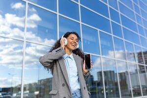 giovane bellissimo latino americano donna passeggiate nel il città, donna nel cuffie ascolta per musica e in linea Radio flusso, Saluti e canta lungo, detiene Telefono nel mani, usi app. foto