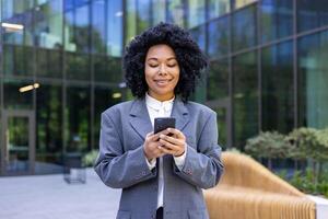 riuscito africano americano attività commerciale donna a piedi nel il città a partire dal al di fuori ufficio costruzione, finanziario lavoratore sorridente con soddisfazione, donna Tenere smartphone utilizzando app, navigazione sociale reti. foto