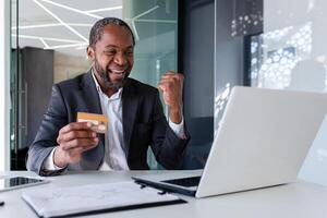 contento giovane africano americano uomo seduta nel ufficio nel davanti di il computer portatile e felice, Tenere credito carta e mostrando vittoria sì gesto. foto