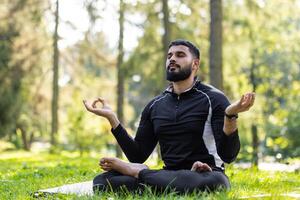 giovane musulmano uomo fare yoga nel il parco. seduta su un' stuoia nel il loto posizione e Meditare con chiuso occhi. foto