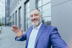 anziano riuscito e esperto grigio dai capelli uomo guardare in Telefono telecamera e sorridente parlando su chiamata, uomo d'affari assunzione autoscatto mentre in piedi al di fuori ufficio edificio foto