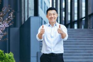 contento asiatico agente uomo d'affari guardare a telecamera e sorridente Tenere pollici su al di fuori ufficio foto