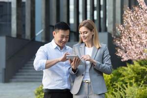 Due lavoratori, asiatico uomo e attività commerciale donna, al di fuori il ufficio, sorridente e felice, guardare a il progetto implementazione Piano nel il tavoletta computer foto