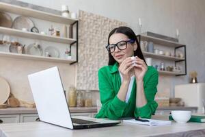 un' giovane donna nel bicchieri e verde Abiti è seduta a casa nel il cucina a il tavolo Guardando un' film, serie, programma a partire dal un' il computer portatile. lei è pianto, Tenere un' tovagliolo nel il suo mani, asciugandosi il suo lacrime. foto