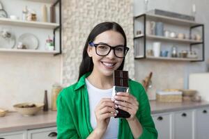 giovane bellissimo donna nel bicchieri e un' verde camicia a casa nel il cucina, Tenere cioccolato nel sua mani, mangiare un' bar di nero cioccolato. Guarda a il telecamera, Sorridi. foto