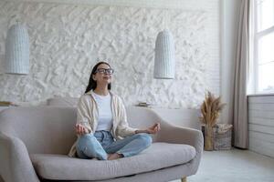 tempo per yoga. attraente giovane donna indossare bicchieri seduta su divano a casa nel loto posizione con chiuso occhi. riposa, rilassa. foto