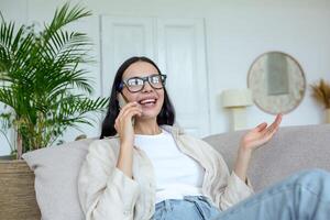 contento giovane bellissimo donna nel bicchieri seduta su il divano a casa e parlando su il Telefono con amici e famiglia. Esprimere a gesti con mani. quarantena, a distanza in linea comunicazione. foto