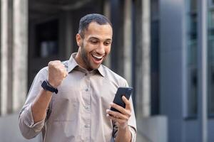 africano americano uomo d'affari esprimendo gioia mentre guardare a il suo smartphone, festeggiare al di fuori un ufficio costruzione. foto