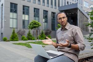 confuso uomo d'affari con il computer portatile all'aperto, esprimendo incertezza, moderno ufficio sfondo, risoluzione dei problemi concetto, a distanza opera. foto