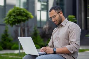 focalizzata uomo Lavorando su il computer portatile all'aperto, scrittura Appunti. concetto di a distanza opera, tecnologia, e concentrazione nel un' casuale ambientazione. foto