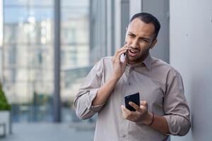 confuso giovane uomo nel attività commerciale casuale abbigliamento parlando su Telefono e guardare a smartphone schermo con un' perplesso espressione. foto