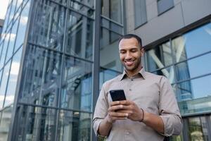 giovane riuscito uomo d'affari al di fuori ufficio edificio utilizzando Telefono, africano americano uomo con smartphone nel mani sfoglia sociale reti, quadranti Messaggio chiamata. foto