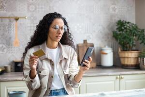 frustrato imbrogliato donna a casa nel cucina respinto, sbagliato i soldi trasferimento, Internet frode, latino americano donna triste Tenere banca credito addebito carta e Telefono, utilizzando in linea shopping app. foto