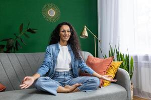 giovane bellissimo donna Meditare nel loto posa, ispanico donna sorridente contento e guardare a telecamera, seduta su divano nel vivente camera a casa, praticante yoga classe. foto