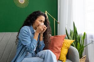 donna nel denim camicia sensazione male, starnuti in fazzoletto di carta su divano, casa interno con impianti, esprimendo malattia e allergia sintomi. foto