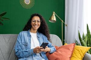 sorridente donna con bicchieri seduta su divano a casa, Tenere Telefono. accogliente, elegante interno con verde parete e impianti. foto