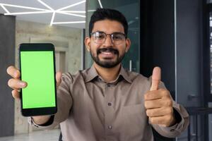 avvicinamento ritratto di un' giovane indiano uomo indossare bicchieri seduta nel il ufficio, mostrando il super cartello con il suo dito per il telecamera e un' verde Telefono schermo modello. foto