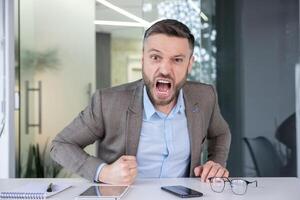 un arrabbiato maschio ufficio lavoratore urlando con stretto pugni, mostrando forte emozione e posto di lavoro fatica contro un' aziendale interno sfondo. foto