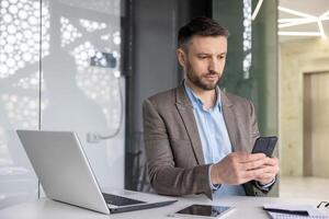 grave concentrato maturo uomo d'affari pensiero lettura in linea sociale reti, esperto maturo uomo Tenere Telefono utilizzando smartphone a posto di lavoro dentro ufficio con il computer portatile. foto