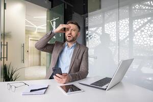 ritratto di un' scioccato giovane uomo seduta nel il ufficio a il scrivania, Tenere il Telefono nel il suo mani e guardare a il telecamera nel sorpresa, Tenere il suo testa. foto