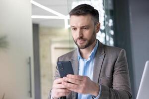 grave concentrato maturo uomo d'affari pensiero lettura in linea sociale reti, esperto maturo uomo Tenere Telefono utilizzando smartphone a posto di lavoro dentro ufficio con il computer portatile. foto