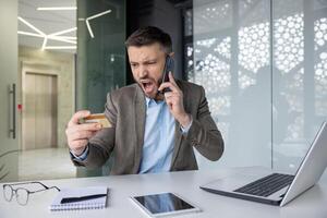 uomo d'affari esprimendo frustrazione su Telefono mentre guardare a credito carta, concetto di finanziario frode e fatica nel un' moderno ufficio ambientazione. foto