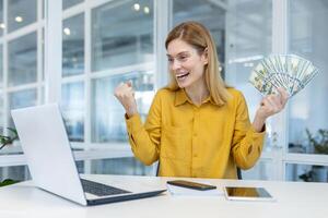 eccitato donna d'affari nel giallo camicia celebra finanziario successo, Tenere denaro contante mentre utilizzando un' il computer portatile nel un' contemporaneo ufficio ambientazione. foto