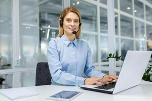 un' professionale donna indossare un' leggero blu camicia e cuffia si siede a un' bianca ufficio scrivania, digitando su un' il computer portatile nel un' luminosa, moderno ufficio ambientazione. foto