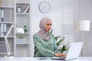 focalizzata musulmano donna nel hijab a opera su sua il computer portatile nel un' ben illuminato, moderno ufficio ambiente con un' pulito design. foto