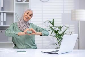 un' allegro giovane musulmano donna indossare un' hijab fa un' cuore forma con sua mani, esprimendo positività e amore nel un' luminosa, contemporaneo ufficio ambientazione. foto