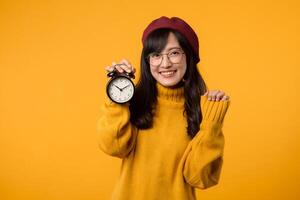 un' allegro giovane asiatico donna, nel un' giallo maglione e rosso berretto, sta Il prossimo per un allarme orologio, simboleggiante puntualità e stile contro un' vivace giallo sfondo. foto