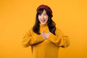 giovane asiatico donna nel sua anni '30, indossare un' giallo maglione e rosso berretto, trasporta gratitudine e gioia con un' commovente mano gesto contro un' giallo sfondo. foto