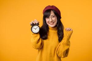 puntualità incontra stile come un' giovane asiatico donna, indossare un' giallo maglione e rosso berretto, lampeggia un' contento Sorridi accanto sua fidato allarme orologio nel sua elegante spazio di lavoro. foto