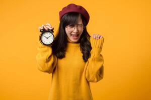 un' giovane asiatico donna con un' contento Sorridi, indossare un' giallo maglione e rosso berretto, sta con fiducia Il prossimo per un allarme orologio nel sua elegante ufficio. foto