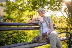 contento anziano donna gode potabile caffè e la spesa tempo con sua gatto mentre seduta su un' panchina nel sua giardino. foto