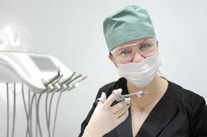 femmina medico nel un' dentale ufficio nel un' maschera e occhiali verde berretto per contenere capelli nero completo da uomo nel sua mani carpale siringa e igienico chirurgico guanti bianca parete ultimo tecnologia attraversare su il il petto foto