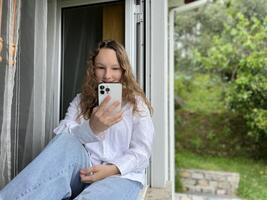 un' ragazza nel un' bianca camicia un' adolescente si siede su un' bianca davanzale il Visualizza a partire dal il strada lei può essere visto nel il finestra nel sua mani lei detiene un' bianca i phone 13 professionista max lei sembra a il schermo con calma foto