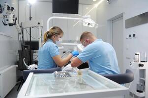 medico trattamento a il dentista ufficio lato Visualizza paziente e preparazione lui per il trattamento. grave uomo nel dentista sedia guardare a il medico e chiede per qualcosa dentista e paziente foto