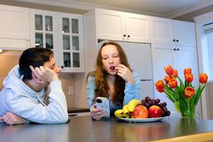 adolescenti nel il cucina un' ragazzo sembra a un' ragazza con amorevole occhi un' giovane donna mangia uva frutta un' uomo Leans su il tavolo e delicatamente esamina sua primo amore mobile Telefono foto