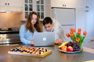 impostato su un' il computer portatile riparazione adolescenti preparato per orologio Sushi su tavolo tulipani frutta nel cucina celebrare marzo 8 San Valentino giorno compleanno film attentamente focalizzata Guarda a aggeggio cucinando vacanza divertimento foto