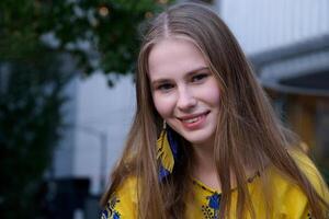 giovane bellissimo donna, giovane ragazza nel grande città nel giallo ricamato camicia, ricamato camicetta, lungo Marrone capelli, Sorridi, piuma orecchini, giallo e blu colori nel Abiti, ucraino nazionale costume foto