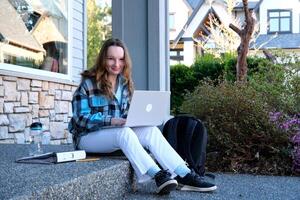 un' adolescenziale ragazza è studiando seduta contro il sfondo di grigio Casa nel il computer portatile e digitando opera Là è cartella e acqua Il prossimo per studia in linea studia in linea opera primo lavoro scuola Università esame foto